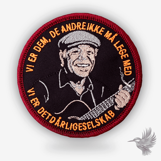 Rundt broderet patch med portræt af Kim Larsen og teksten “Vi er dem de andre ikke må lege med – vi er det dårlige selskab” i orange og rød