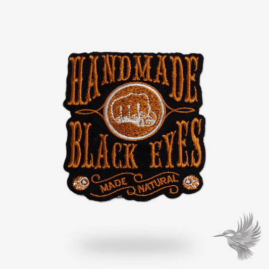 Broderet strygemærke med knytnæve og teksten “Handmade black eyes made natural,” ideelt til at dekorere jakker, tasker eller tøj med en stærk og unik stil.