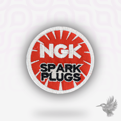 NGK Spark Plugs patch strygemærke med rød baggrund og hvitt logo, symbol på motorsport og mekanisk perfektion.